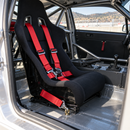 A black racing bucket seat with bright red harness straps installed inside a stripped race car interior with a roll cage and metal floor.