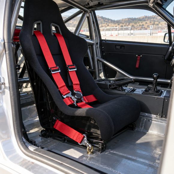 A black racing bucket seat with bright red harness straps installed inside a stripped race car interior with a roll cage and metal floor.